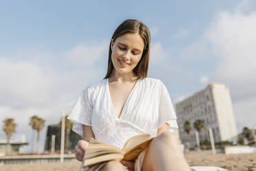 Beautiful young woman reading book while sitting at beach - XLGF02219