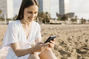 Junge Frau, die am Strand sitzend ein Mobiltelefon benutzt - XLGF02220