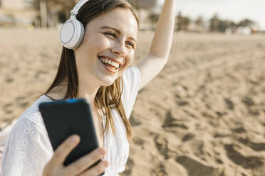 Glückliche Frau, die ein Handy hält und tanzt, während sie über Kopfhörer am Strand Musik hört - XLGF02224