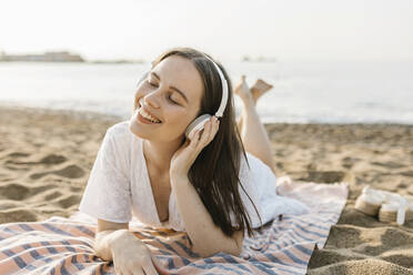 Lächelnde Frau hört Musik über Kopfhörer, während sie auf einem Handtuch liegt - XLGF02225