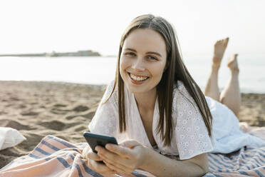 Lächelnde Frau mit Handy in der Hand beim Entspannen am Strand - XLGF02229