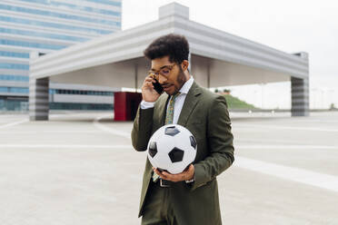 Männlicher Profi, der einen Fußball hält, während er mit einem Mobiltelefon spricht - MEUF04134