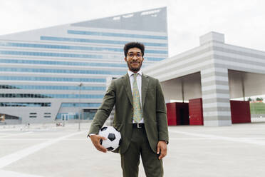 Lächelnder männlicher Profi, der einen Fußball hält, während er in der Stadt steht - MEUF04139