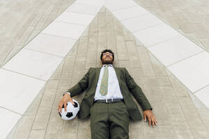 Businessman with soccer ball lying on footpath - MEUF04142