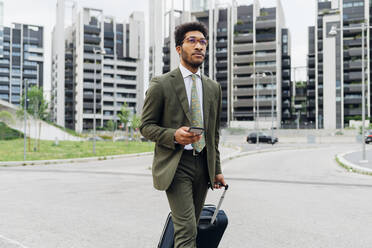Young businessman holding mobile phone and wheeled luggage while walking in city - MEUF04154