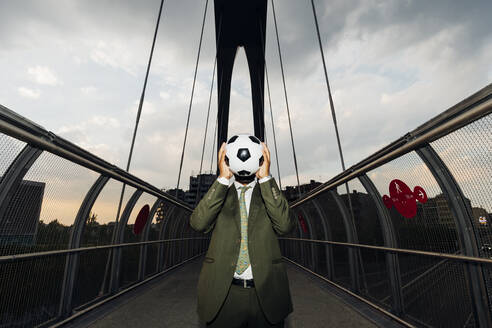 Geschäftsmann, der sein Gesicht mit einem Fußball bedeckt, während er auf einer Brücke steht - MEUF04155