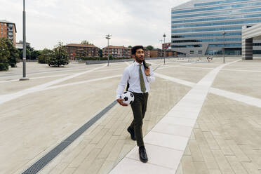 Businessman with soccer ball and blazer walking on footpath - MEUF04165