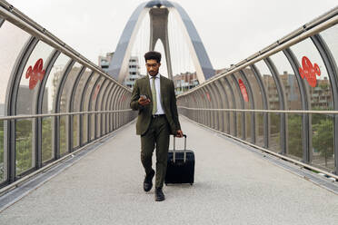 Young businessman using mobile phone while walking with wheeled luggage on bridge - MEUF04168