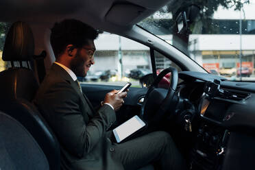 Männlicher Berufstätiger, der ein Mobiltelefon benutzt, während er mit einem digitalen Tablet im Auto sitzt - MEUF04175