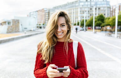 Smiling woman holding smart phone while standing on road - MGOF04735
