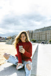 Happy woman using smart phone while sitting on retaining wall - MGOF04738