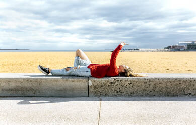 Glückliche Frau, die ihr Smartphone benutzt, während sie am Strand auf einer Stützmauer liegt - MGOF04740