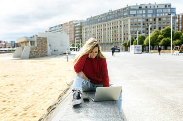 Lächelnde Frau, die einen Laptop benutzt, während sie auf einer Stützmauer am Strand sitzt - MGOF04741