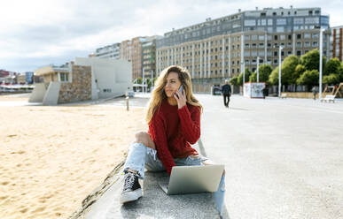 Smiling woman talking on phone while using laptop on retaining wall - MGOF04742