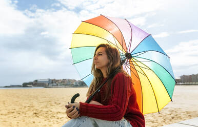 Frau mit Regenschirm, die am Strand sitzt und träumt - MGOF04752