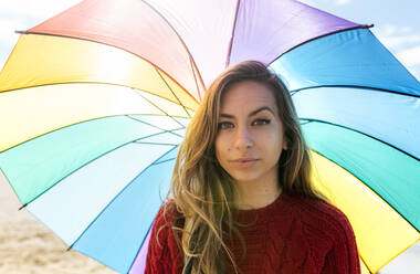 Lächelnde mittelgroße erwachsene Frau mit mehrfarbigem Regenschirm an einem sonnigen Tag - MGOF04753