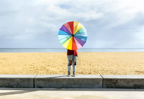 Frau hält Regenschirm, während sie auf der Mauer vor dem Strand steht - MGOF04754