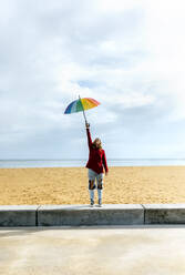 Frau hält Regenschirm mit erhobener Hand, während sie vor einem bewölkten Himmel am Strand steht - MGOF04755