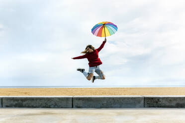 Woman holding umbrella with hand raised while jumping in front of cloudy sky at beach - MGOF04756