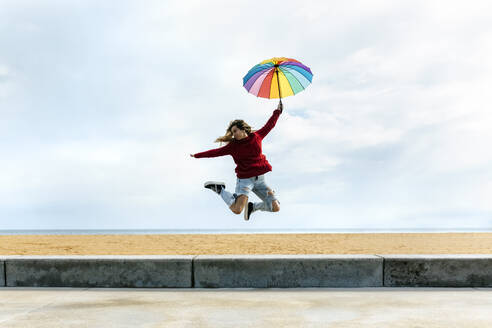 Frau hält Regenschirm mit erhobener Hand, während sie vor dem bewölkten Himmel am Strand springt - MGOF04756