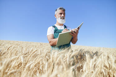 Lächelnder männlicher Landarbeiter, der mit einem Buch auf einem Weizenfeld steht und wegschaut - KIJF04124