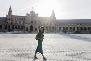 Lächelnde junge Frau, die beim Spaziergang auf der Plaza De Espana, Sevilla, Spanien, wegschaut - JRVF01504