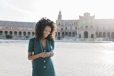 Glückliche junge Frau, die auf der Plaza De Espana, Sevilla, Spanien, eine Textnachricht über ihr Smartphone verschickt - JRVF01506