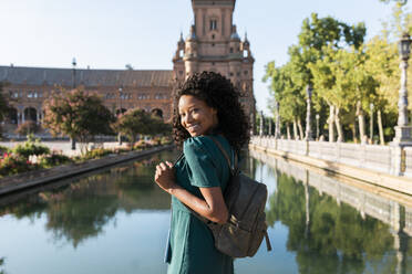 Lächelnde junge Afro-Frau mit Rucksack am Teich auf der Plaza De Espana, Sevilla, Spanien - JRVF01509