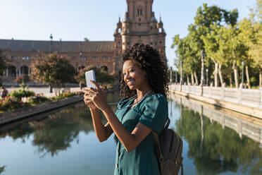 Lächelnde junge Afro-Frau, die am Teich auf der Plaza De Espana, Sevilla, Spanien, ein Mobiltelefon benutzt - JRVF01511