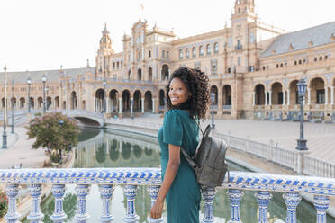 Lächelnde junge Frau mit lockigem Haar an einem Geländer auf der Plaza De Espana, Sevilla, Spanien - JRVF01514