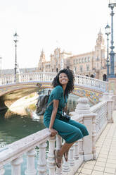 Lächelnde junge Frau mit lockigem Haar, die auf einem Geländer am Plaza De Espana, Sevilla, Spanien, sitzt - JRVF01521