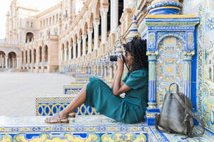 Weibliche Touristin mit lockigem Haar, die durch die Kamera fotografiert, während sie auf der Plaza De Espana sitzt, Sevilla, Spanien - JRVF01525
