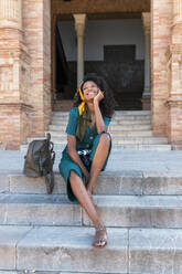 Lächelnde Frau, die mit der Hand am Kinn auf der Plaza De Espana, Sevilla, Spanien, sitzt und träumt - JRVF01529