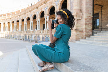 Lächelnde Touristin mit lockigem Haar, die eine Kamera hält, während sie auf den Stufen der Plaza De Espana, Sevilla, Spanien, sitzt - JRVF01530