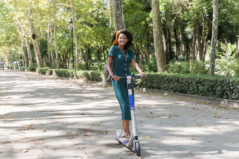 Glückliche junge Frau, die wegschaut, während sie einen elektrischen Roller im Park fährt - JRVF01542