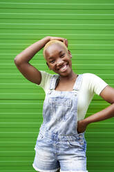 Glückliche junge Frau steht mit der Hand auf der Hüfte vor einer grünen Wand - VEGF04871