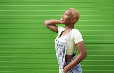 Lächelnde junge Frau blickt auf, während sie mit der Hand in der Tasche an einer grünen Wand steht - VEGF04873