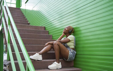Junge Frau träumt, während sie über drahtlose Kopfhörer auf einer Treppe Musik hört - VEGF04881