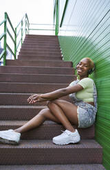Lächelnde junge Frau, die auf einer Treppe sitzend über Kopfhörer Musik hört - VEGF04882