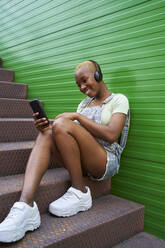 Smiling woman using smart phone while listening music at staircase - VEGF04883