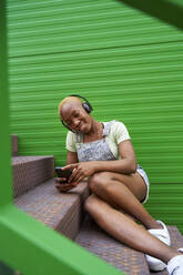 Young woman listening music through headphones while using mobile phone on staircase - VEGF04885