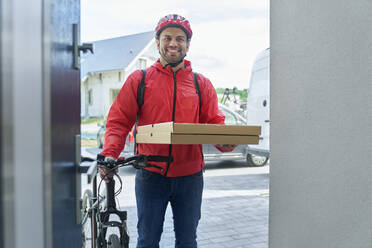 Ein lächelnder Pizzabote hält einen Pizzakarton in der Hand, während er mit einem Fahrrad vor der Tür steht - ABIF01455