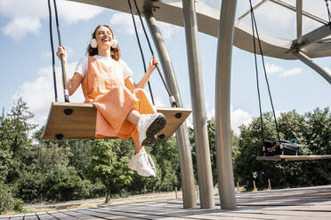 Fröhliche Frau hört Musik über Kopfhörer, während sie im Park schaukelt - VPIF04573