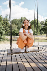 Smiling woman with headphones crouching on swing at park - VPIF04574