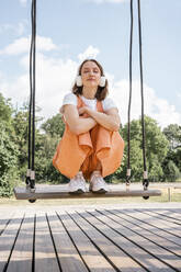 Young woman crouching with arms crossed on swing at park - VPIF04575