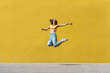 Cheerful woman with arms outstretched jumping on footpath - VPIF04603