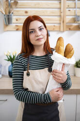 Junge Frau hält einen Laib Brot in der Hand und steht in der Küche - GIOF13217