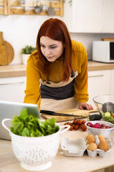 Redhead woman using laptop while preparing food in kitchen - GIOF13232