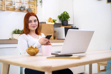 Smiling redhead woman with mobile phone and laptop sitting at home - GIOF13236