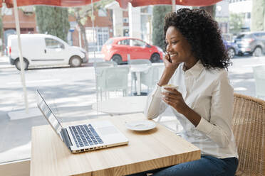 Female freelancer holding coffee cup while listening to video call through laptop at cafe - JRVF01566